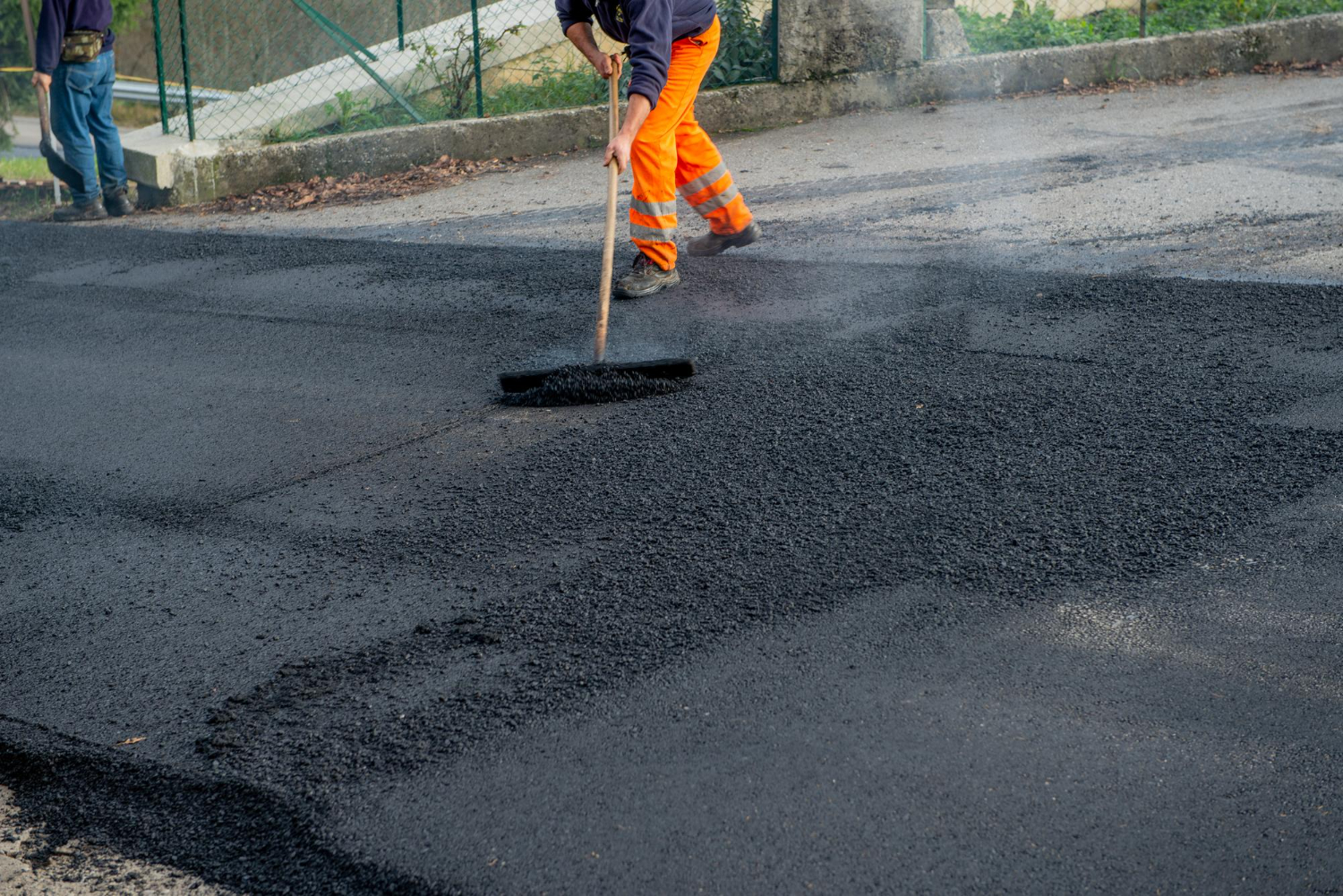 Worker in orange pants spreads hot asphalt on a road with a rake, smoothing the surface.