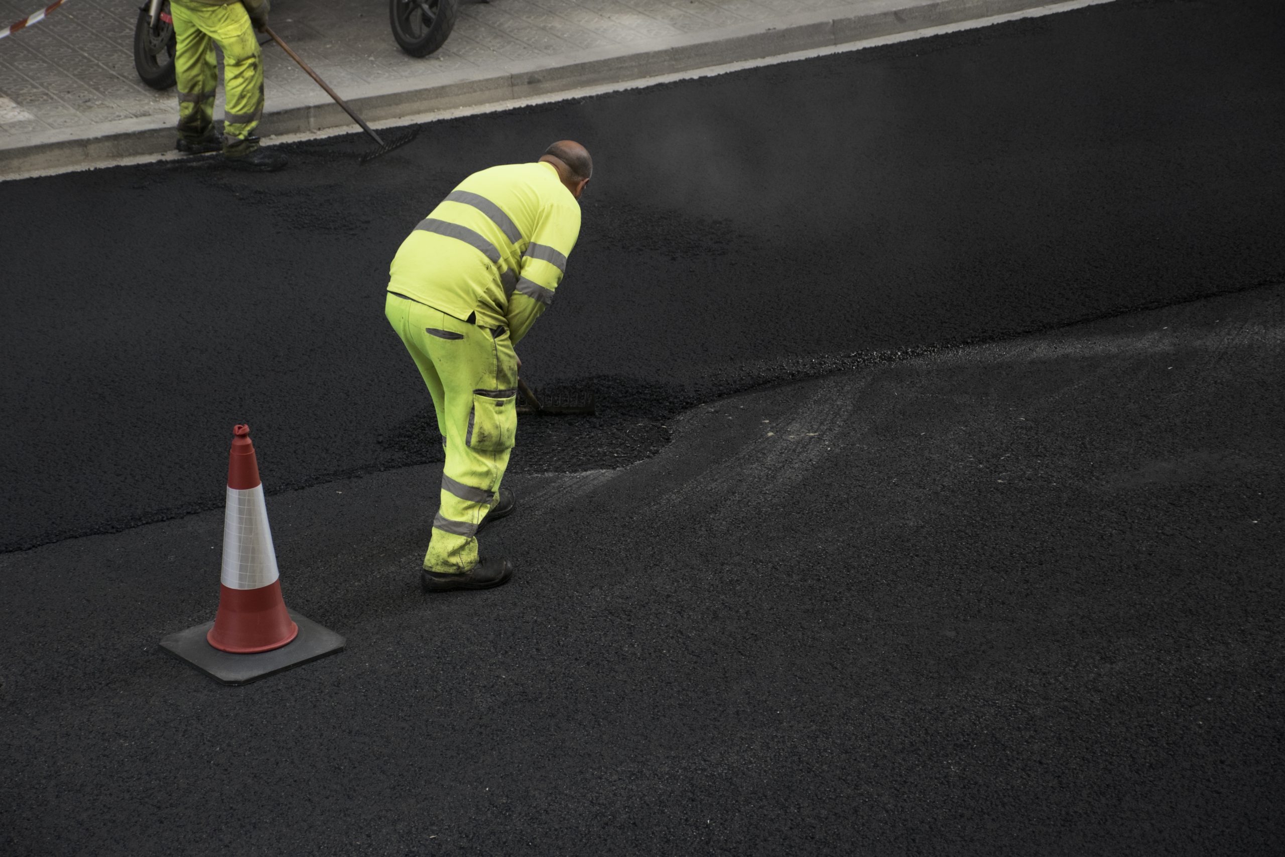 A worker in a yellow uniform smooths fresh asphalt on a road next to an orange traffic cone.