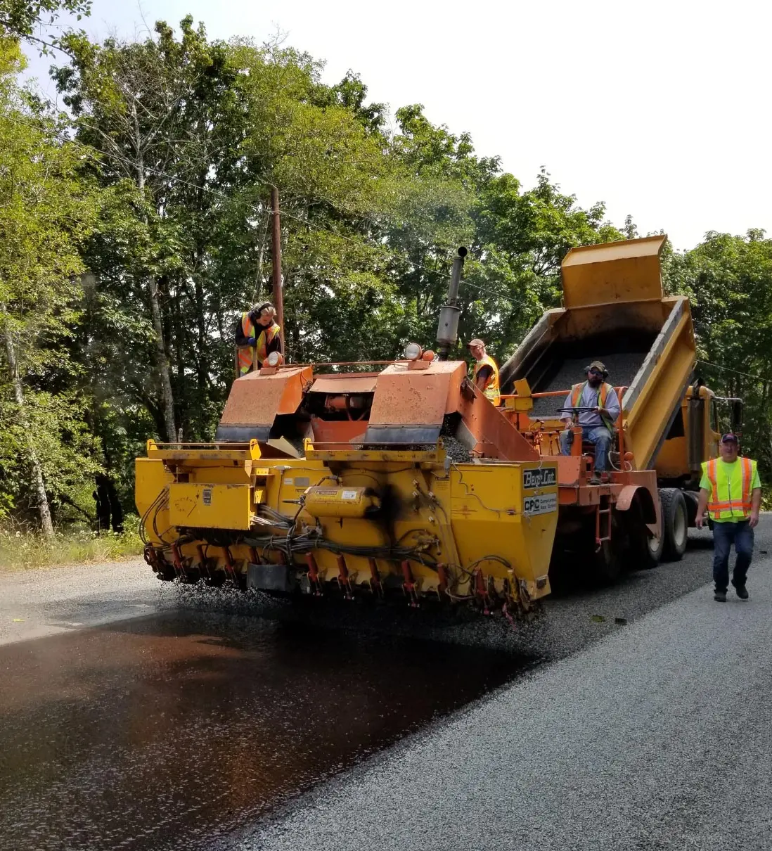 Workers operate machinery laying gravel and tar on a road, with trees and greenery in the background.